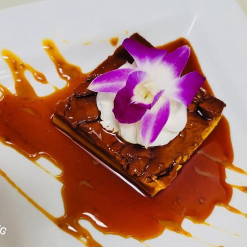 A square dessert with caramel sauce, whipped cream, and a purple orchid on a white plate.