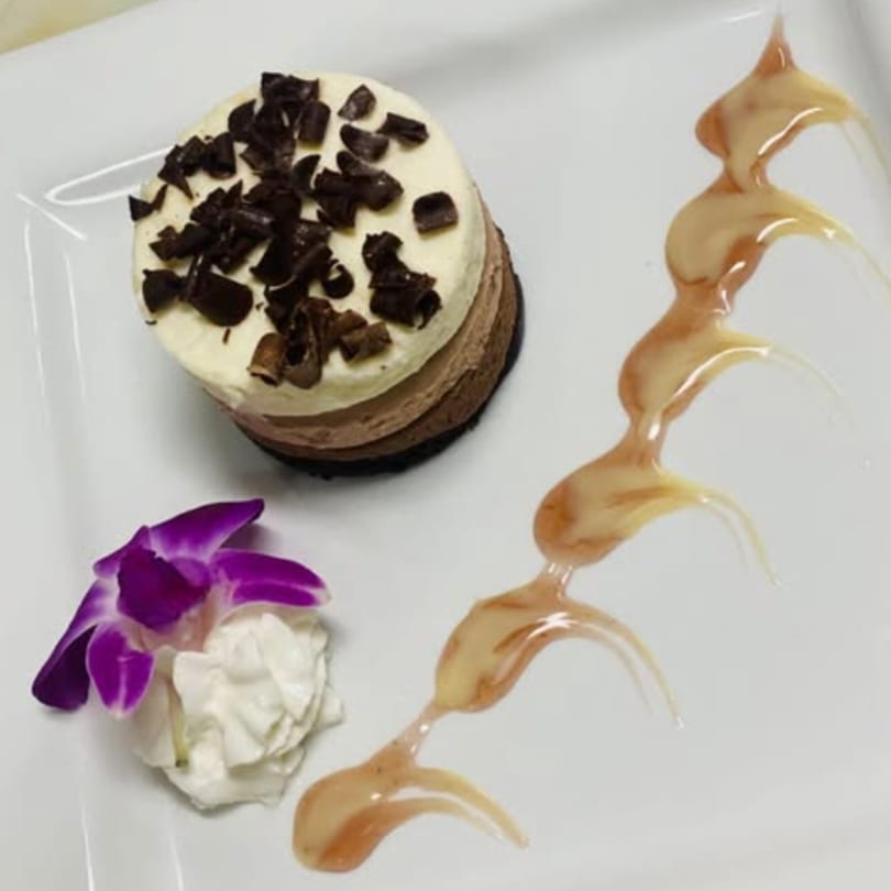 A layered chocolate dessert with chocolate shavings, whipped cream, a purple orchid, and caramel drizzle.